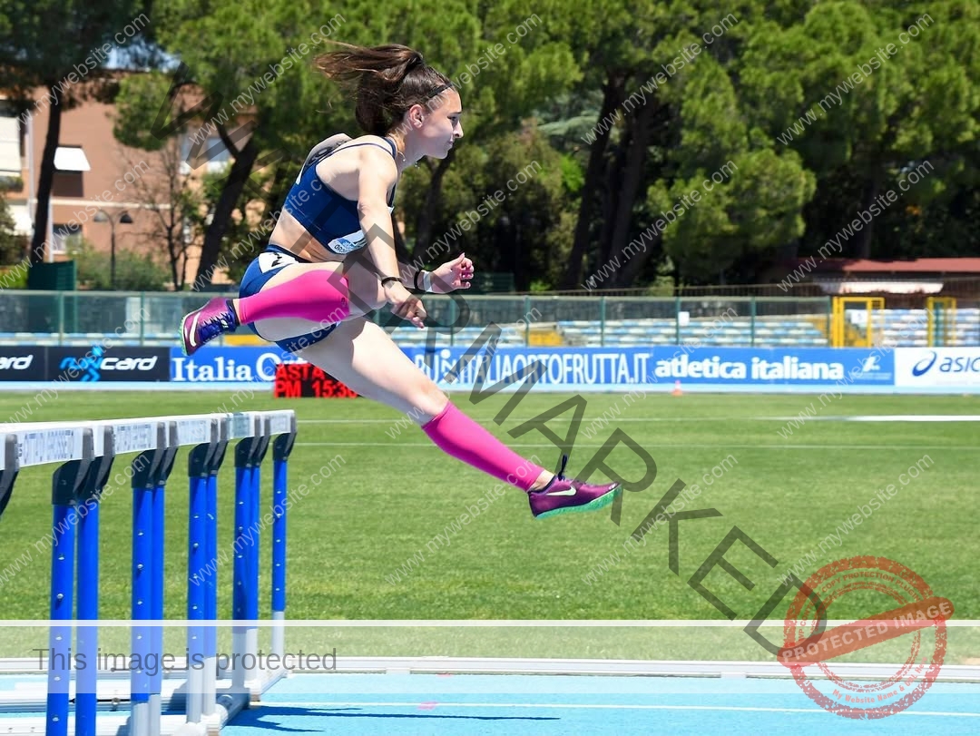 anabel-vitale-italy-anabelvitale-0407 Anabel Vitale, track and field star from Italy, in pink socks leaps over a hurdle on a sunny track with trees and banners behind.
