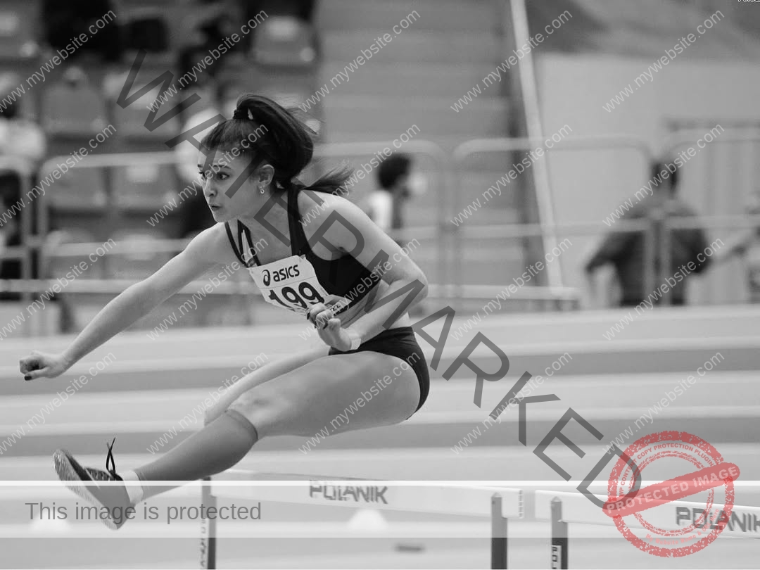 anabel-vitale-italy-anabelvitale-0429 Anabel Vitale, track and field star from Italy, clears a hurdle mid-jump in a black and white indoor track event, focused and determined.