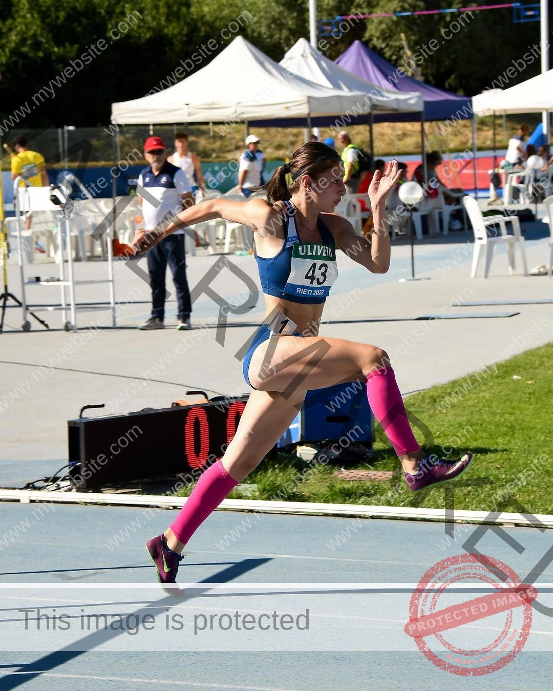 anabel-vitale-italy-anabelvitale-0439 Anabel Vitale, track and field star from Italy, is mid-air in a long jump wearing blue and pink at an outdoor event.