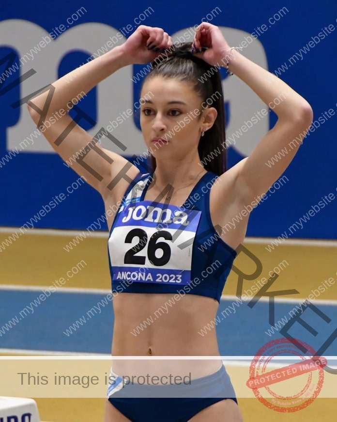 Anabel Vitale, track and field star from Italy, in blue gear with bib 26, adjusts her ponytail before a race on an indoor track.