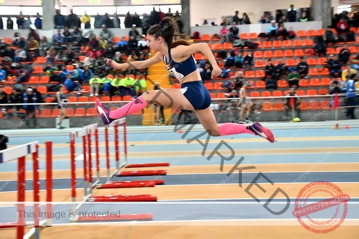 anabel-vitale-italy-anabelvitale-0536 Anabel Vitale, track and field star from Italy, leaps over a hurdle in blue sportswear and pink socks on an indoor track with spectators.