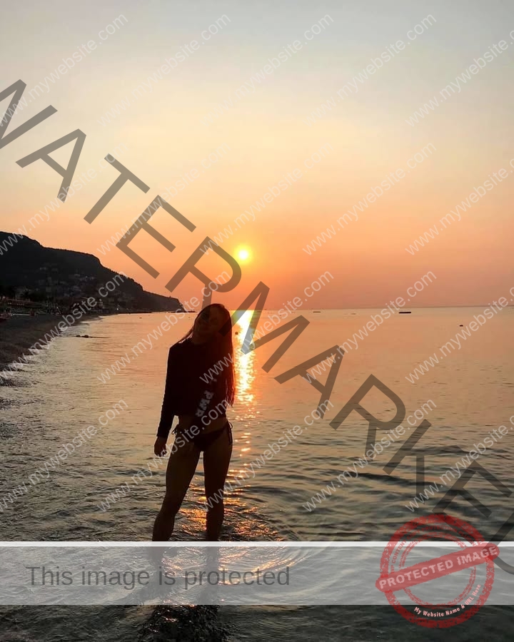 anabel-vitale-italy-anabelvitale-0544 Anabel Vitale, track and field star from Italy, stands in shallow water at sunset, orange sun low over calm sea and distant town.