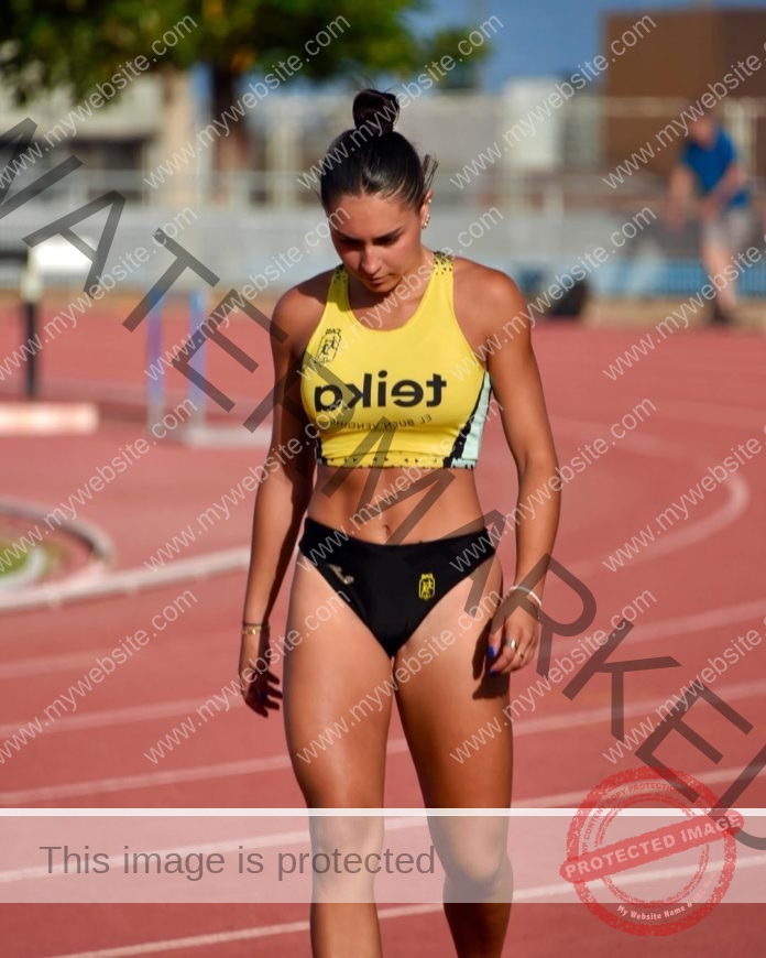 Camilla Pagani, track and field star from Spain, walks on a running track in yellow crop top and black shorts, looking down. Hurdles visible.