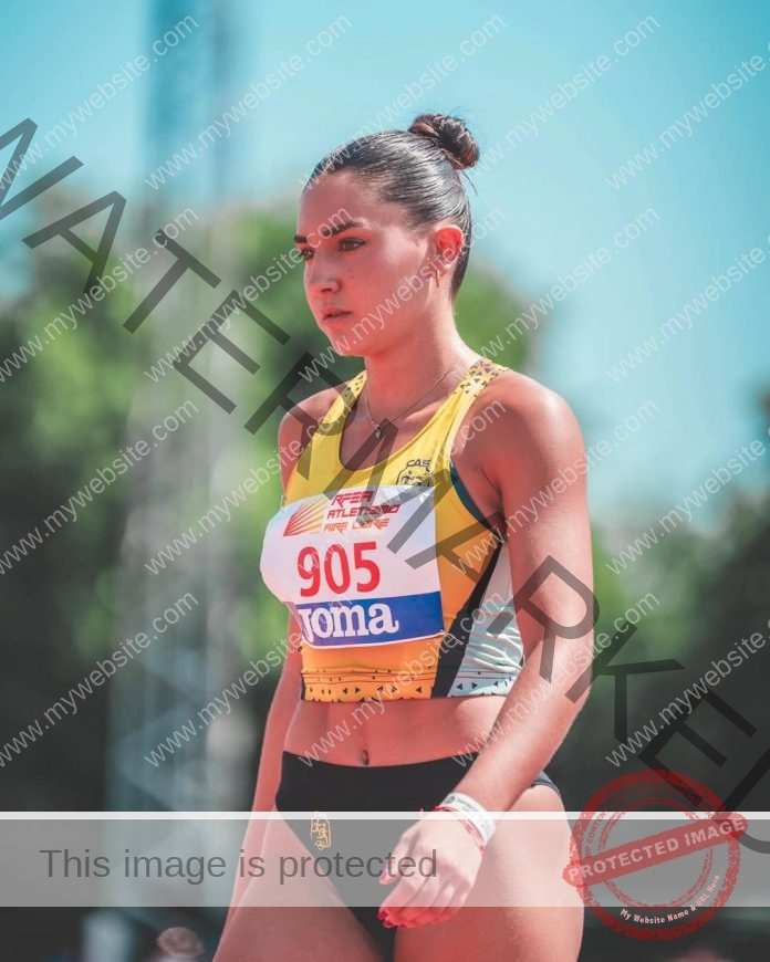 Camilla Pagani, track and field star from Spain, in a yellow sports top and black shorts with bib 905, stands focused outdoors.