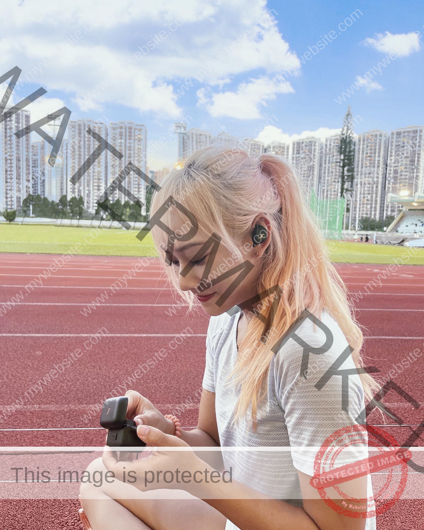 chan-pui-kei-hong-kong-chanpuikeii-02855 Chan Pui Kei, track and field star from Hong Kong, sits on a running track smiling with a small case and earbud; city buildings behind.