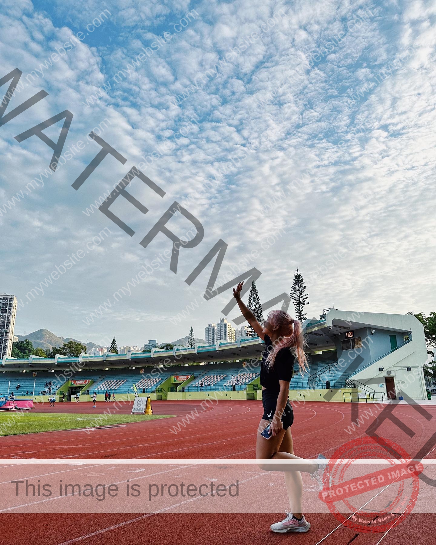 chan-pui-kei-hong-kong-chanpuikeii-02868 Chan Pui Kei, track and field star from Hong Kong, with pink hair poses on a red track; stadium seats and buildings behind her.