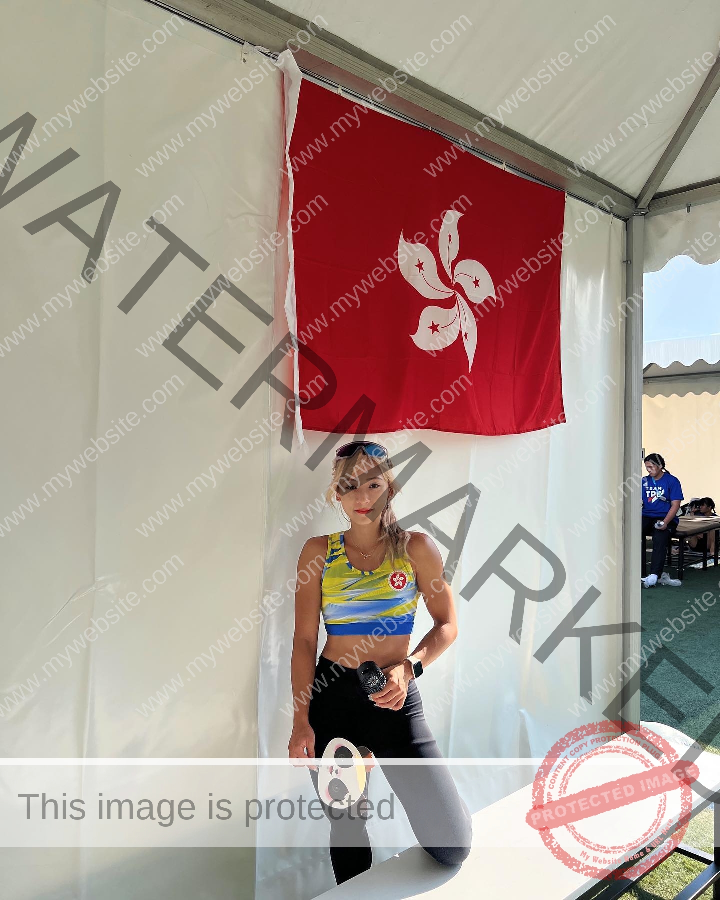 chan-pui-kei-hong-kong-chanpuikeii-02935 Chan Pui Kei, track and field star from Hong Kong, stands under a flag in a tent, paddle and water bottle in hand, serious expression.