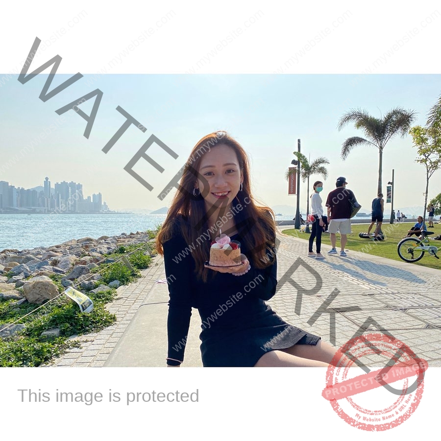 chan-pui-kei-hong-kong-chanpuikeii-03105 Chan Pui Kei, track and field star from Hong Kong sits by the water in a black dress, smiling with a pink cake as people walk and cycle nearby.