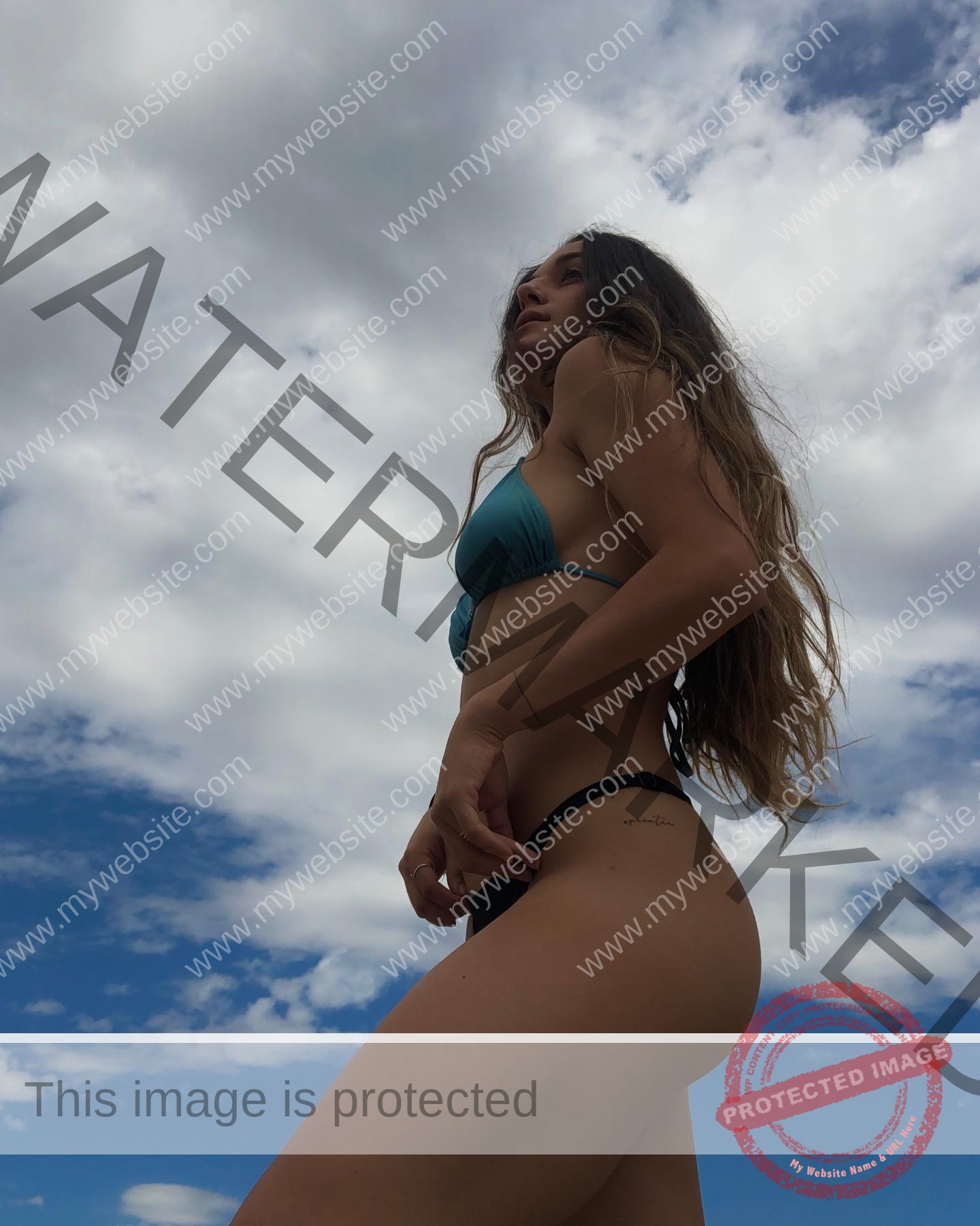 claudia-camacho-spain-clauudiacamacho-1390 Claudia Camacho, track and field star from Spain, stands outdoors in a blue bikini under a cloudy sky, looking upward from below.