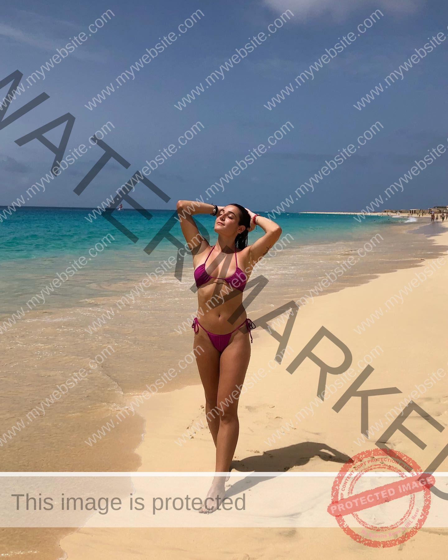 claudia-camacho-spain-clauudiacamacho-1394 Claudia Camacho, track and field star from Spain, in a pink bikini poses on an empty beach by clear blue water under partly cloudy skies.