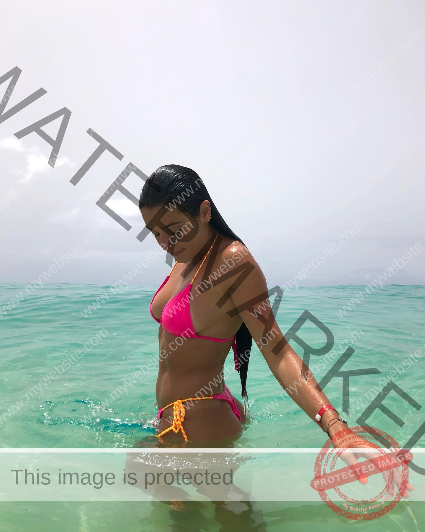 claudia-camacho-spain-clauudiacamacho-1398 Claudia Camacho, track and field star from Spain, stands in turquoise water under clouds, wearing a pink bikini, looking down.