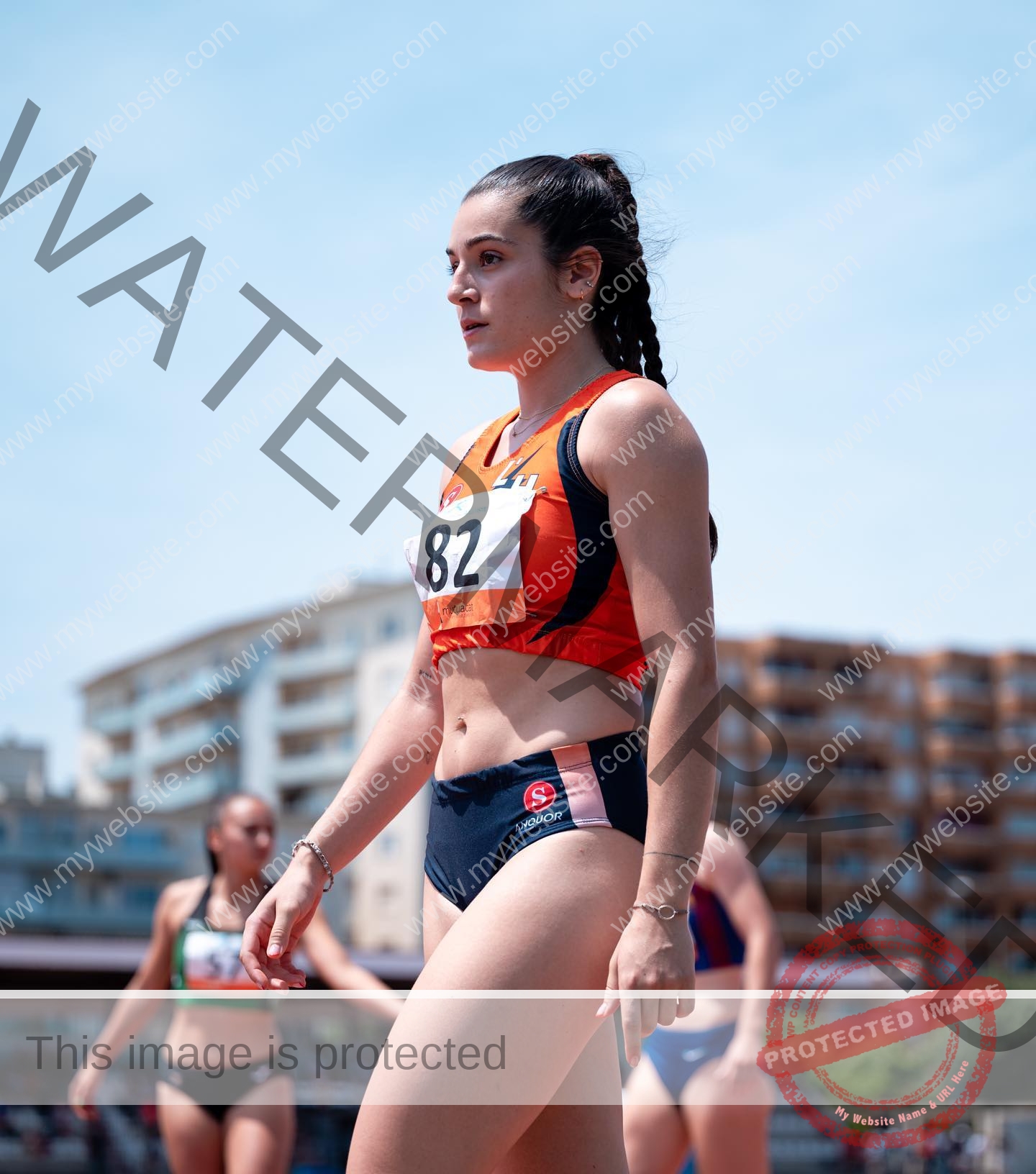claudia-camacho-spain-clauudiacamacho-1405 Claudia Camacho, track and field star from Spain, stands confidently on the track in orange and navy with number 82, background blurred.