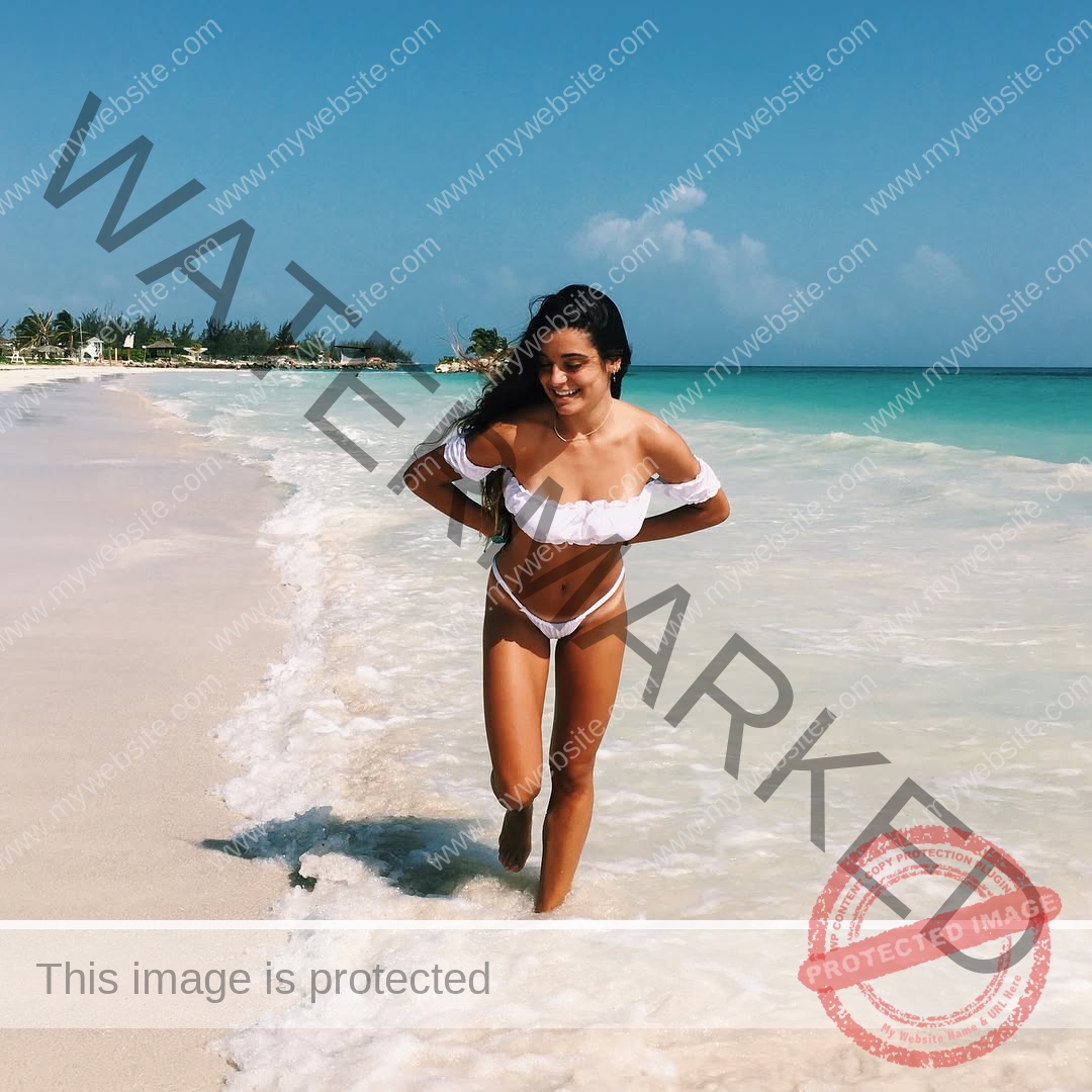 claudia-camacho-spain-clauudiacamacho-1421 Claudia Camacho, track and field star from Spain, smiles in a white bikini walking along the sunny shoreline by clear blue water.