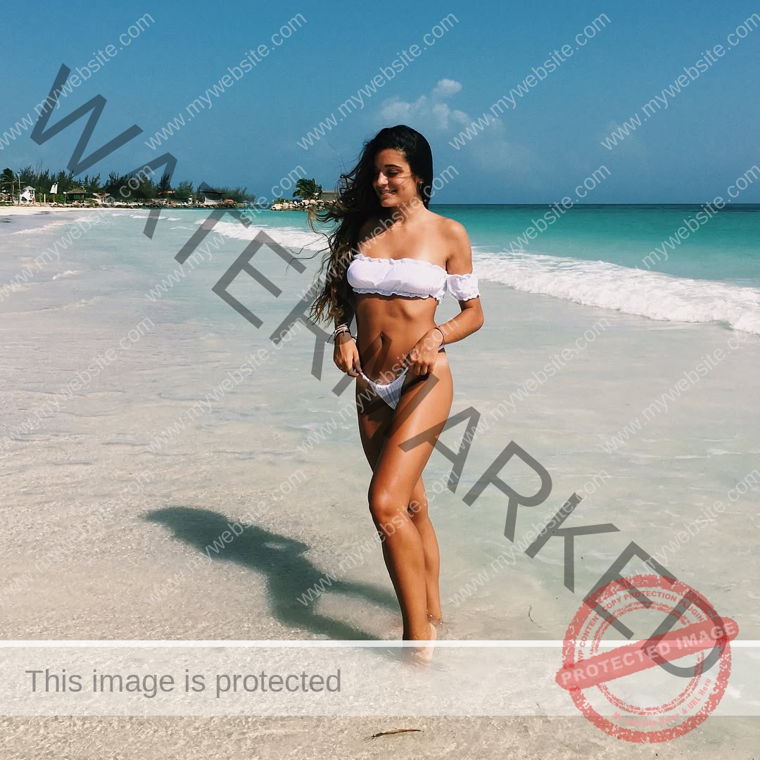 claudia-camacho-spain-clauudiacamacho-1425 Claudia Camacho, track and field star from Spain, in a white bikini walking along a tropical beach, smiling under the bright sky.