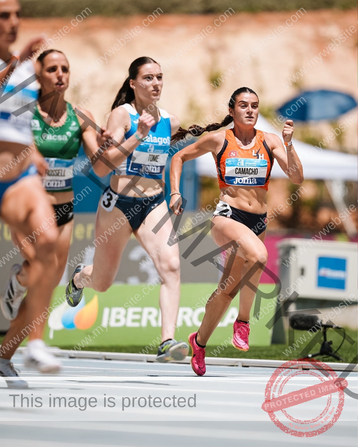 claudia-camacho-spain-clauudiacamacho-1438 Claudia Camacho, track and field star from Spain runs on a track with three others; she leads in orange and black, crowd blurred behind.