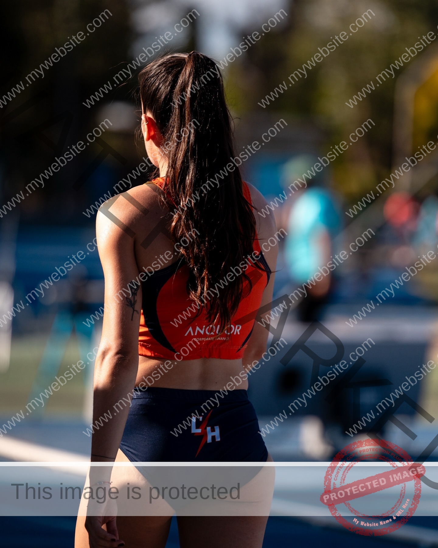 claudia-camacho-spain-clauudiacamacho-1440 Claudia Camacho, track and field star from Spain, stands on an outdoor track in orange top and blue shorts, sunlit from behind.