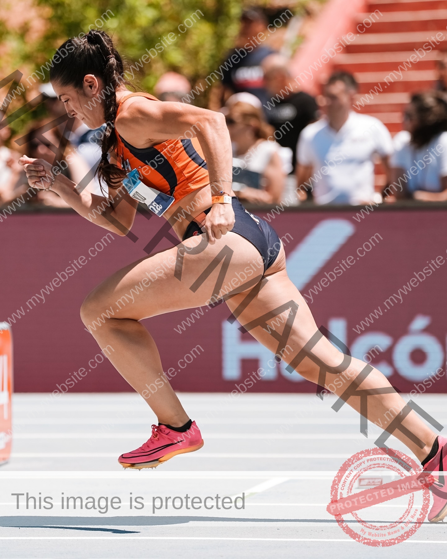 claudia-camacho-spain-clauudiacamacho-1447 Claudia Camacho, track and field star from Spain, sprints in orange and navy on an outdoor track with spectators and a red staircase.