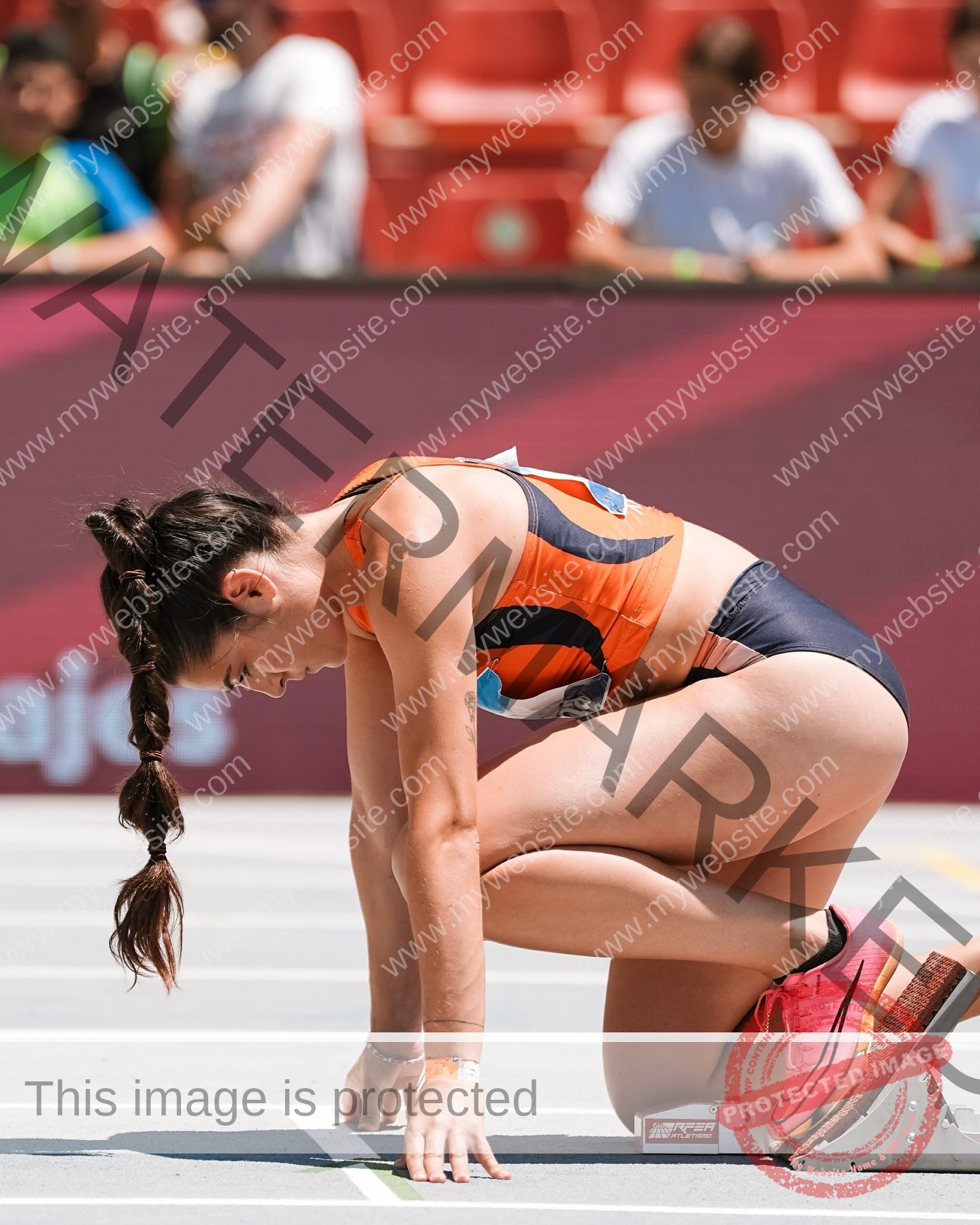 claudia-camacho-spain-clauudiacamacho-1448 Claudia Camacho, track and field star from Spain, kneels at the starting blocks in an orange and navy uniform, ready to race.
