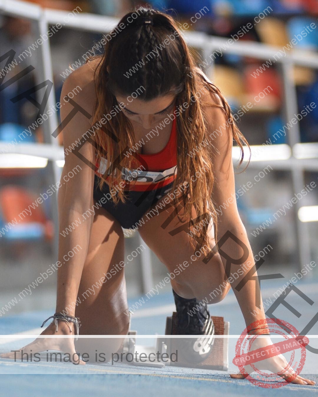claudia-camacho-spain-clauudiacamacho-1463 Claudia Camacho, track and field star from Spain, kneels at the starting blocks in red top and black shorts on a stadium track.