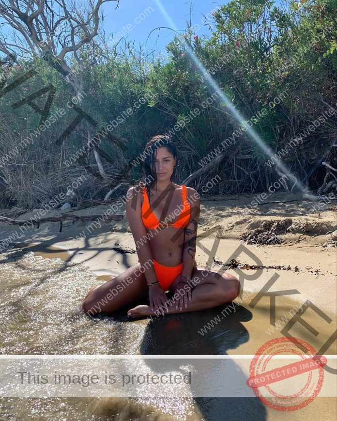 Danai Xiafra, track and field star from Greece, in an orange bikini sits on a sunny sandy shore near water, greenery behind her.