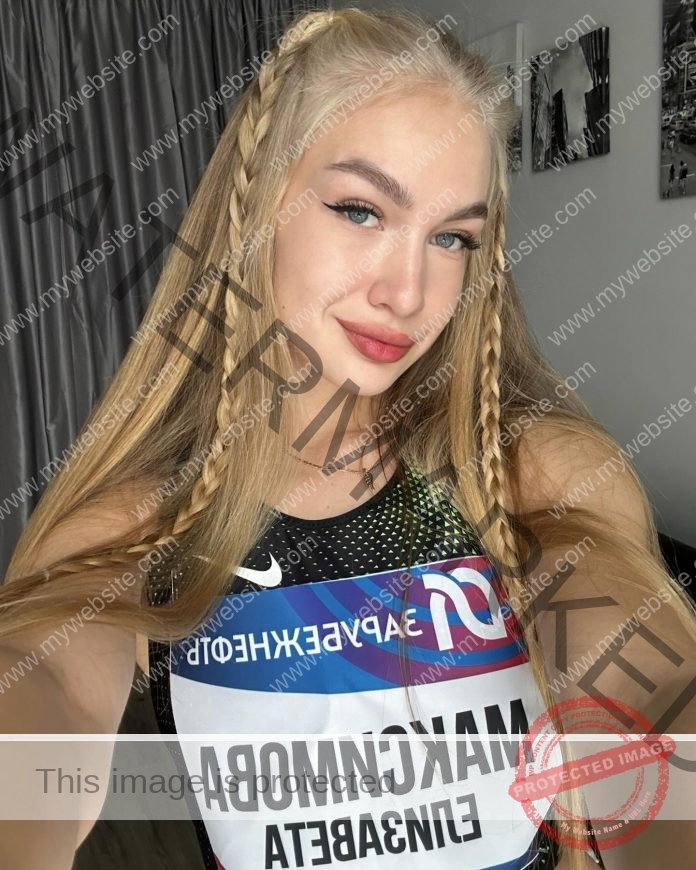 Elizaveta Maksimova, track and field star from Russia, with long blonde braids, smiles in a selfie wearing a Cyrillic sports bib indoors.