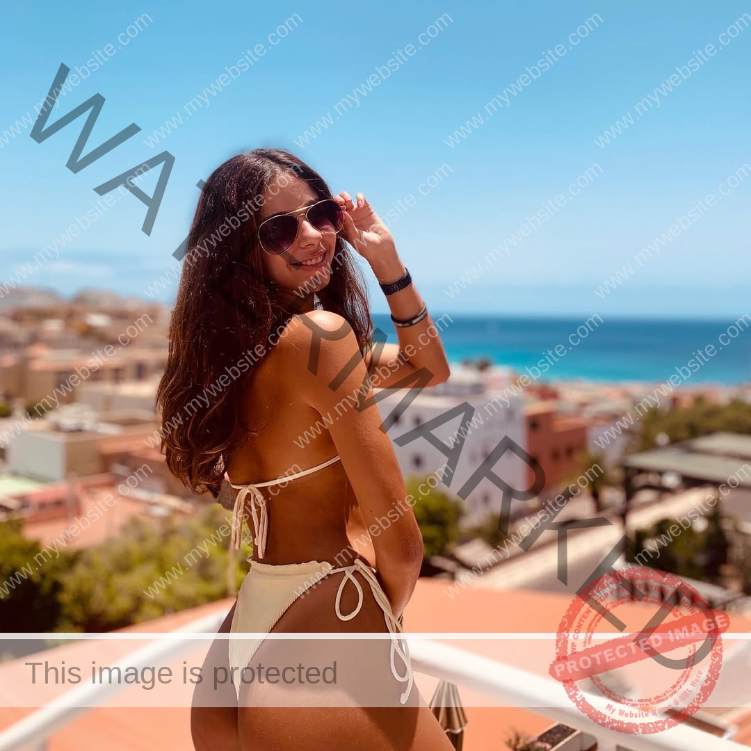 emma-deana-spain-emmadeana-15392 Emma Deana, track and field star from Spain, wears sunglasses and a white bikini on a balcony, smiling with ocean views behind her.