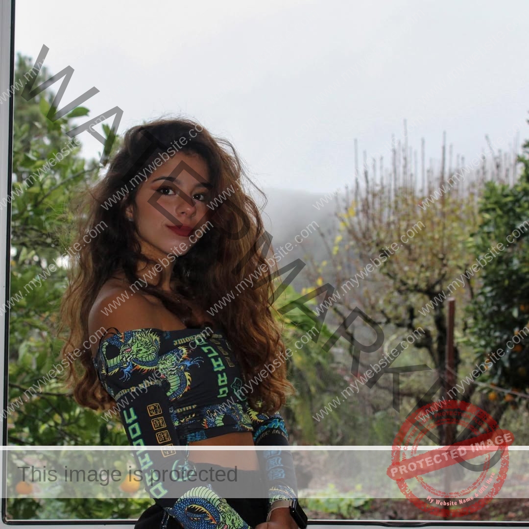 emma-deana-spain-emmadeana-15394 Emma Deana, track and field star from Spain, stands by a window indoors in a colorful off-shoulder top; foggy greenery outside.