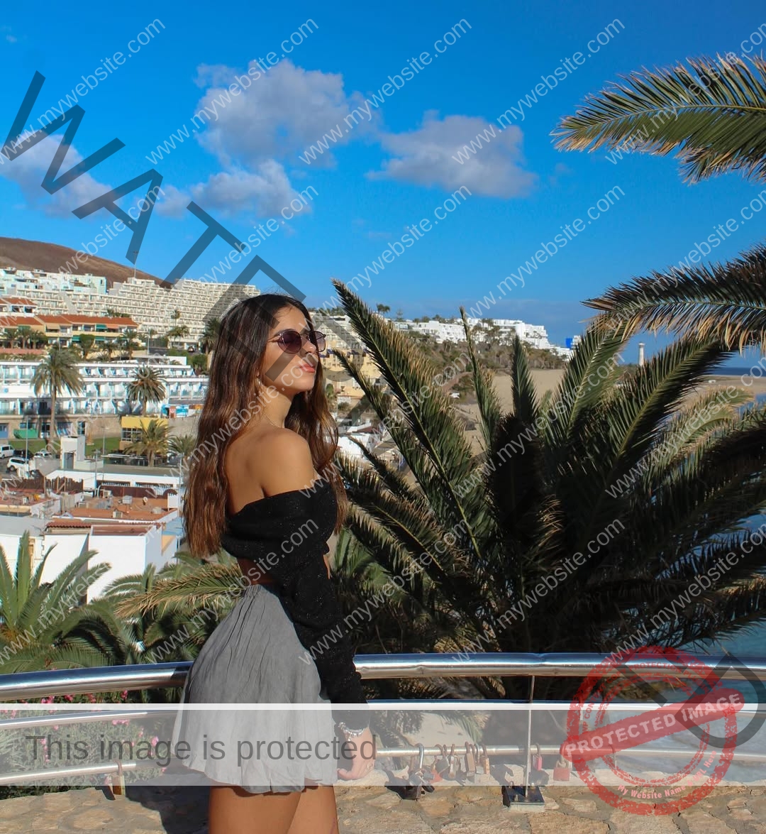 emma-deana-spain-emmadeana-15395 Emma Deana, track and field star from Spain, with long brown hair and sunglasses, poses by a railing under blue sky and palm trees.
