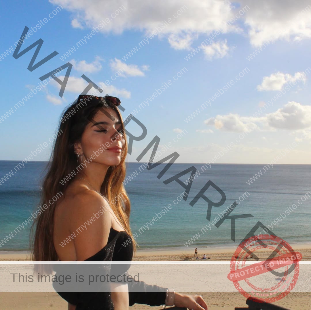 emma-deana-spain-emmadeana-15397 Emma Deana, track and field star from Spain, stands on a beach with eyes closed, smiling in sunlight; ocean and sky behind her.