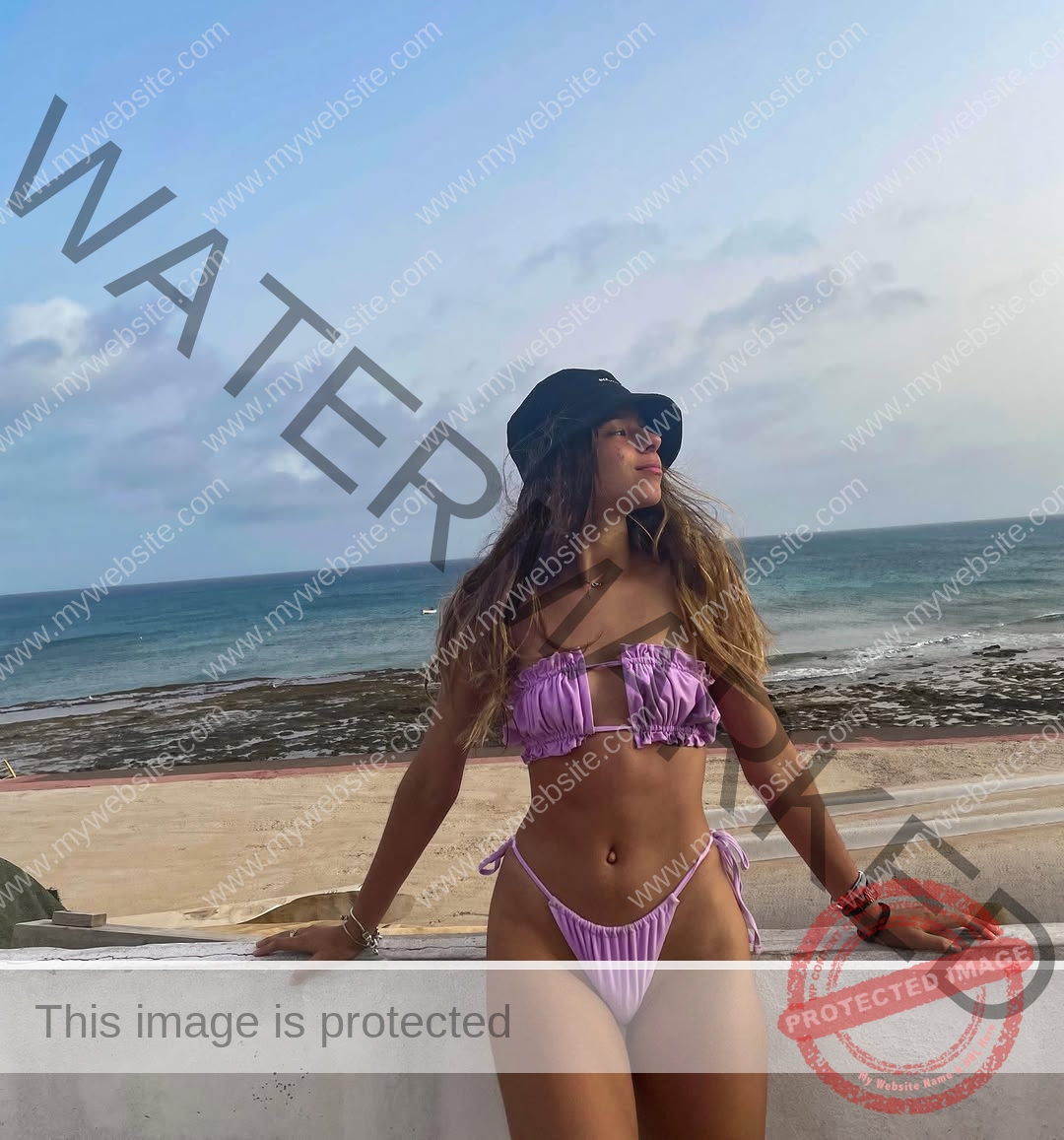 emma-deana-spain-emmadeana-15398 Emma Deana, track and field star from Spain, stands by a white wall in a lavender bikini and black hat, gazing at the beach and ocean.