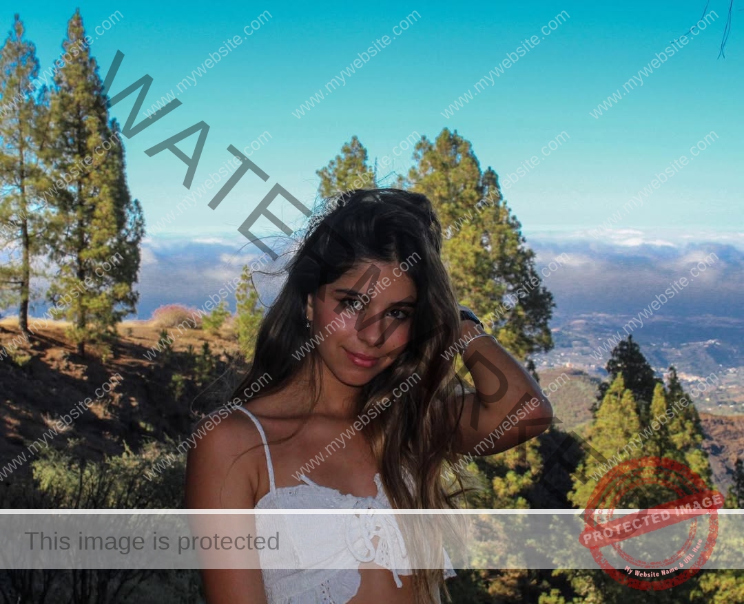 emma-deana-spain-emmadeana-15401 Emma Deana, track and field star from Spain, poses outdoors with one hand in her hair, white sleeveless top, mountains behind.