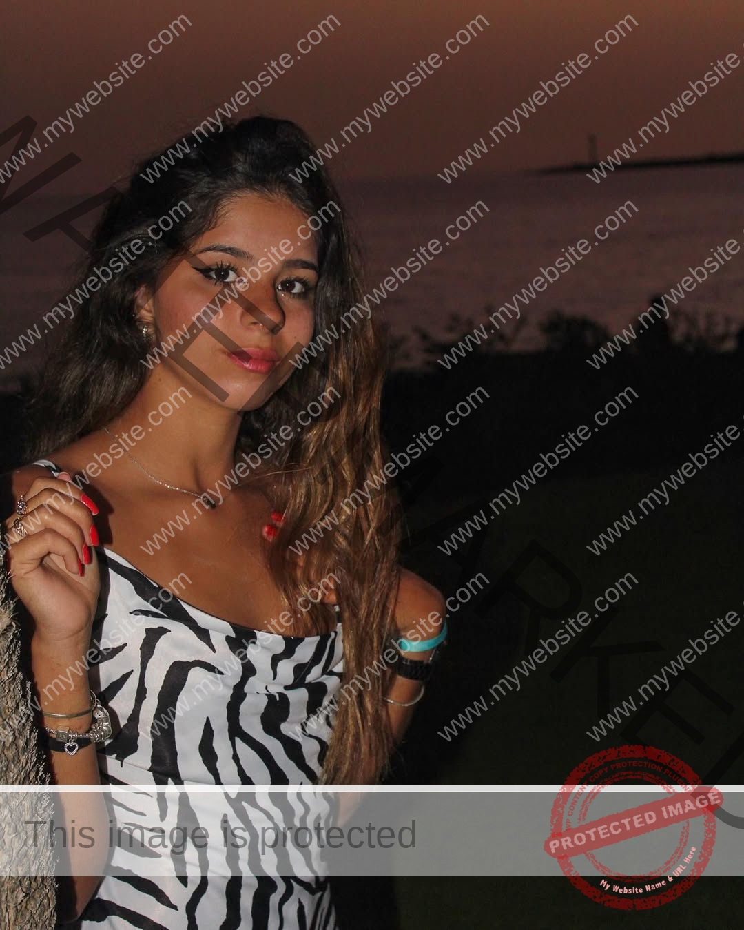 emma-deana-spain-emmadeana-15403 Emma Deana, track and field star from Spain, stands at dusk near water in a zebra-print dress with a beige bag by a reddish sky.