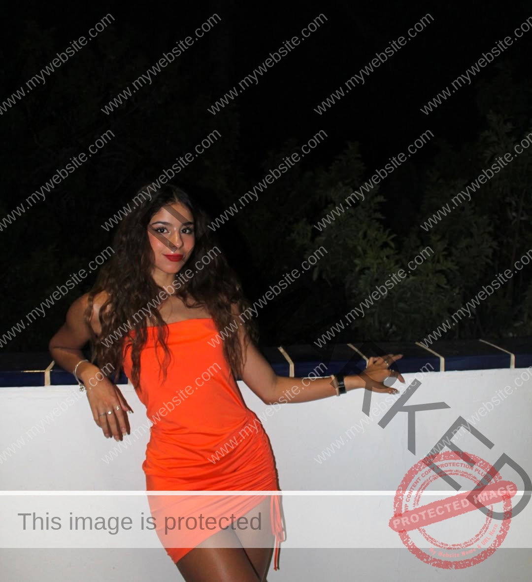 emma-deana-spain-emmadeana-15409 Emma Deana, track and field star from Spain, in an orange dress smiles, leaning on a white wall outdoors at night with foliage behind.