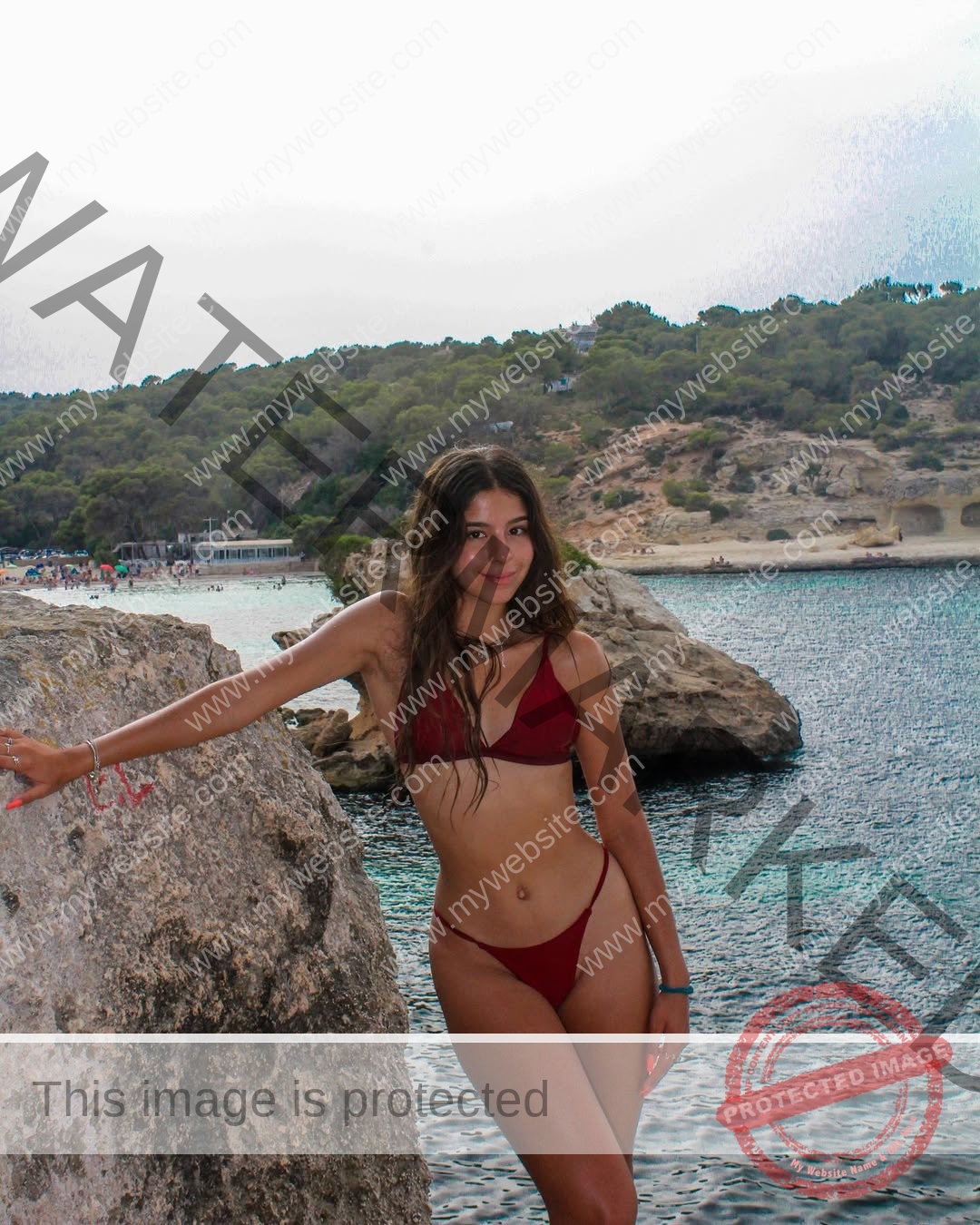 emma-deana-spain-emmadeana-15411 Emma Deana, track and field star from Spain, stands on a rocky shore in a red bikini by the sea with green hills and clouds behind.