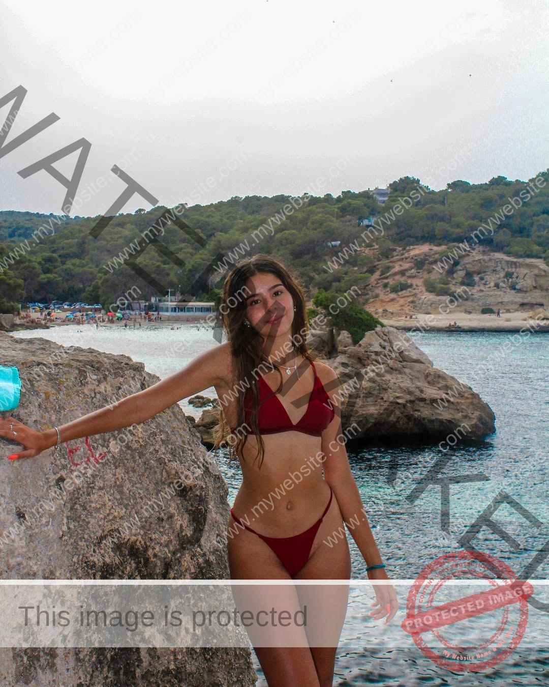 emma-deana-spain-emmadeana-15412 Emma Deana, track and field star from Spain, stands in a maroon bikini on rocky shore, hand on a rock, smiling at the camera.