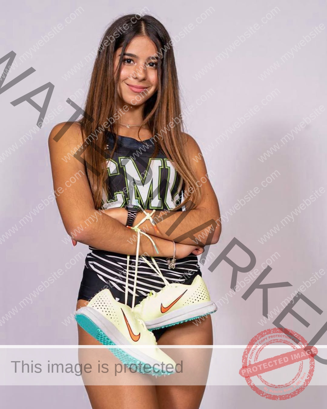 emma-deana-spain-emmadeana-15414 Emma Deana, track and field star from Spain, stands smiling in a CMU uniform, arms crossed, holding white running spikes.