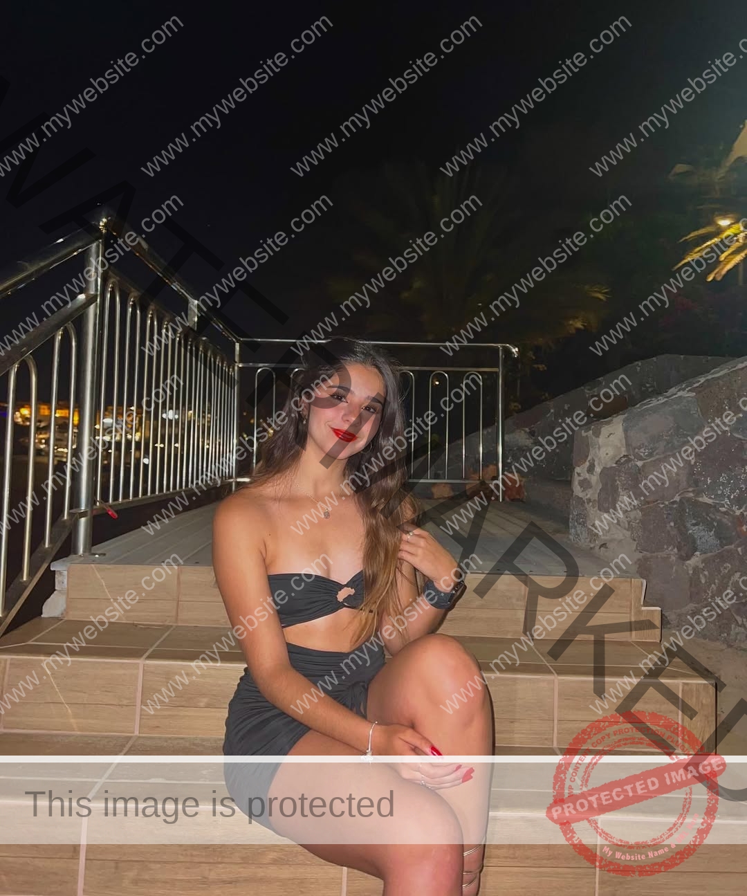 emma-deana-spain-emmadeana-15418 Emma Deana, track and field star from Spain, sits on outdoor stairs at night, smiling in a black strapless top and skirt.