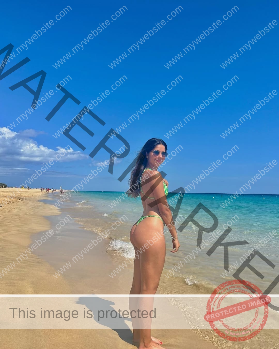 emma-deana-spain-emmadeana-15420 Emma Deana, track and field star from Spain, in a green bikini on a sandy beach, smiles near calm turquoise sea under blue sky.