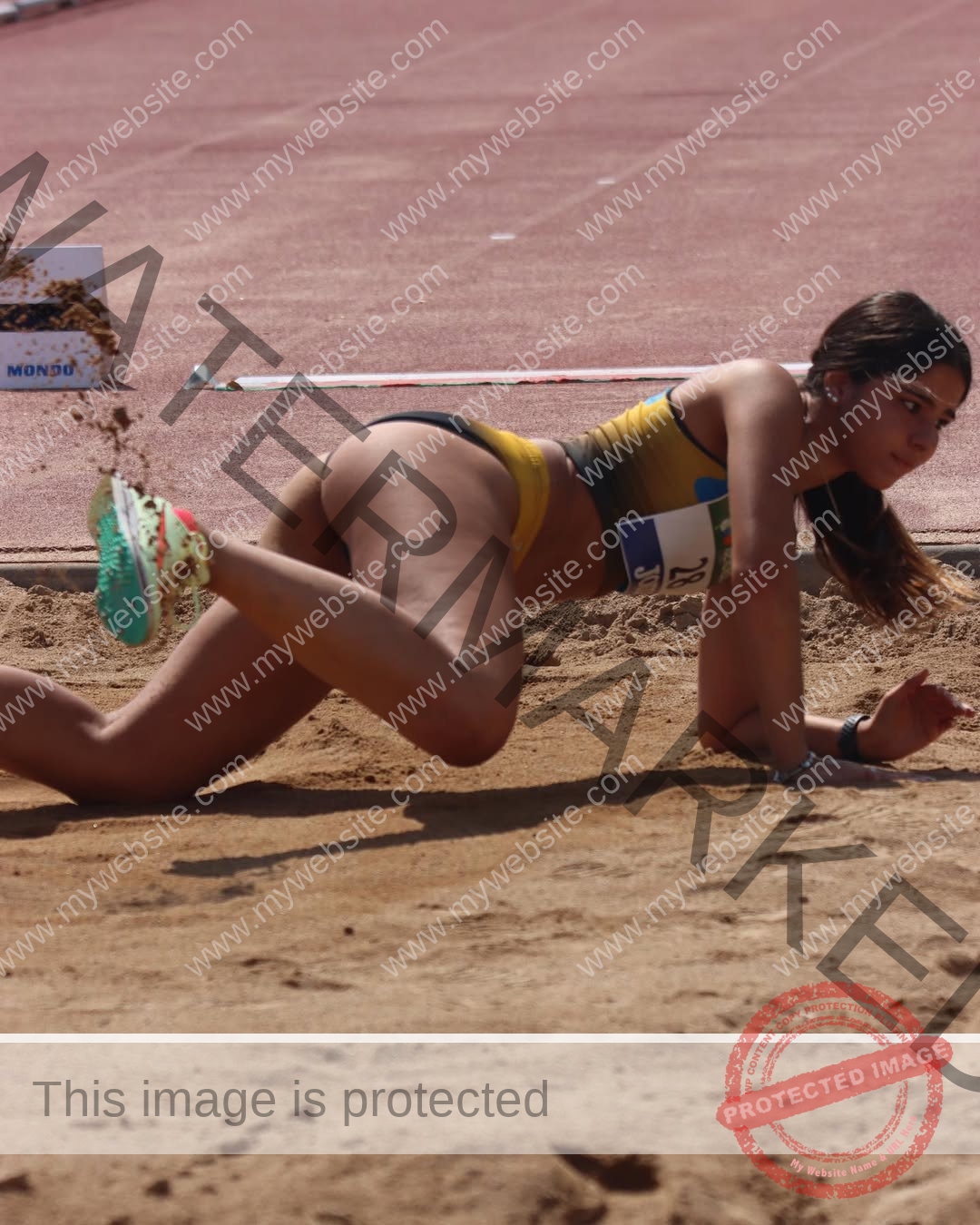 emma-deana-spain-emmadeana-15422 Emma Deana, track and field star from Spain, lands in a sandpit mid-long jump, yellow kit vivid, sand flying as the track blurs behind.