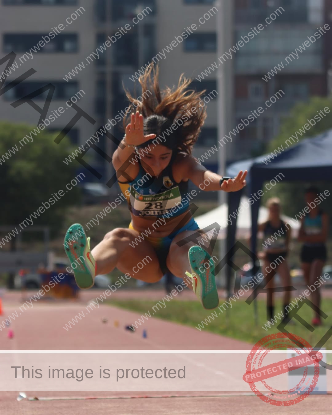 emma-deana-spain-emmadeana-15423 Emma Deana, track and field star from Spain, mid-air in a long jump on an outdoor track, wearing blue and yellow, arms forward.