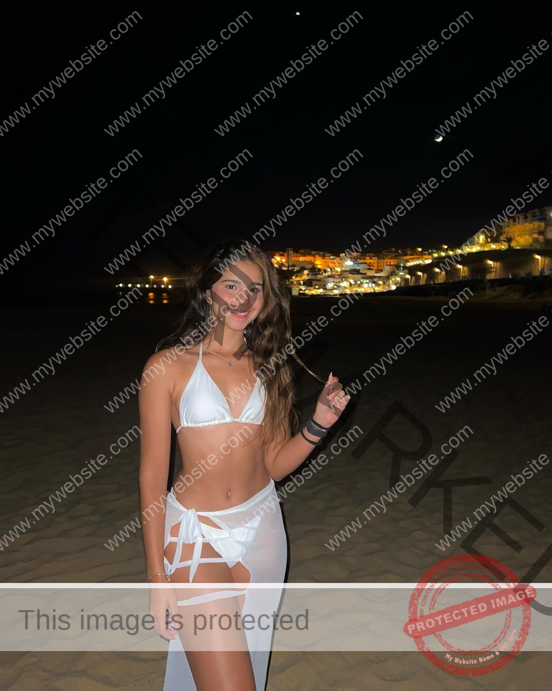 emma-deana-spain-emmadeana-15425 Emma Deana, track and field star from Spain, in a white bikini on a night beach with city lights and crescent moon behind her.