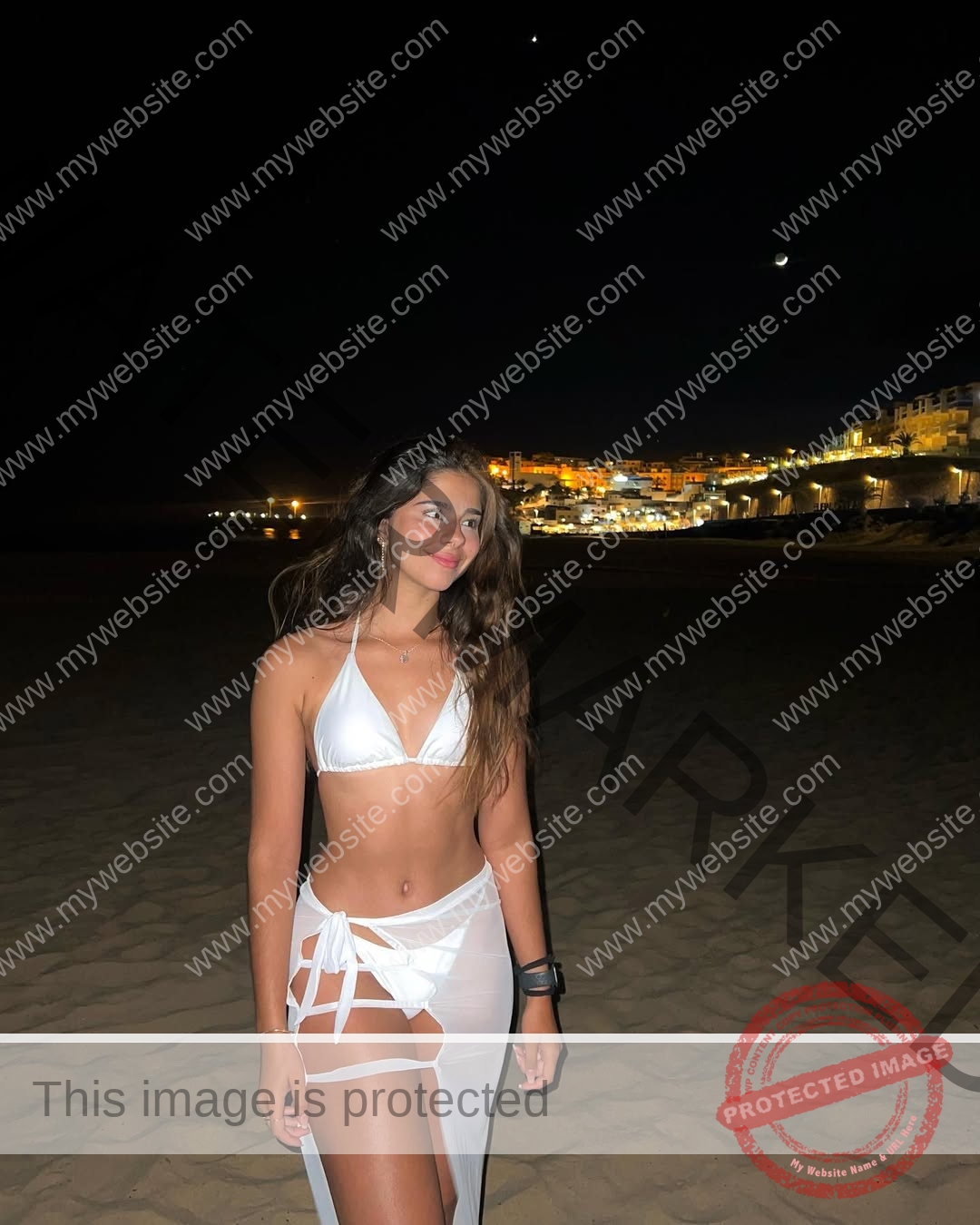 emma-deana-spain-emmadeana-15426 Emma Deana, track and field star from Spain, in a white bikini and sheer wrap smiles on a moonlit beach; city lights behind her.