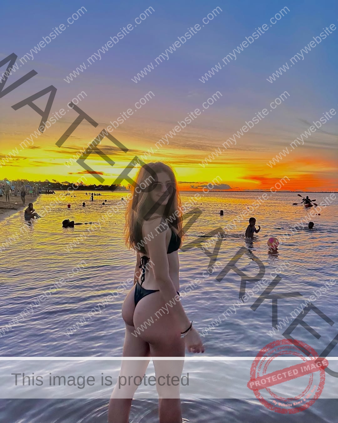 emma-deana-spain-emmadeana-15428 Emma Deana, track and field star from Spain, stands in a black bikini in shallow water, smiling at sunset with swimmers behind her.