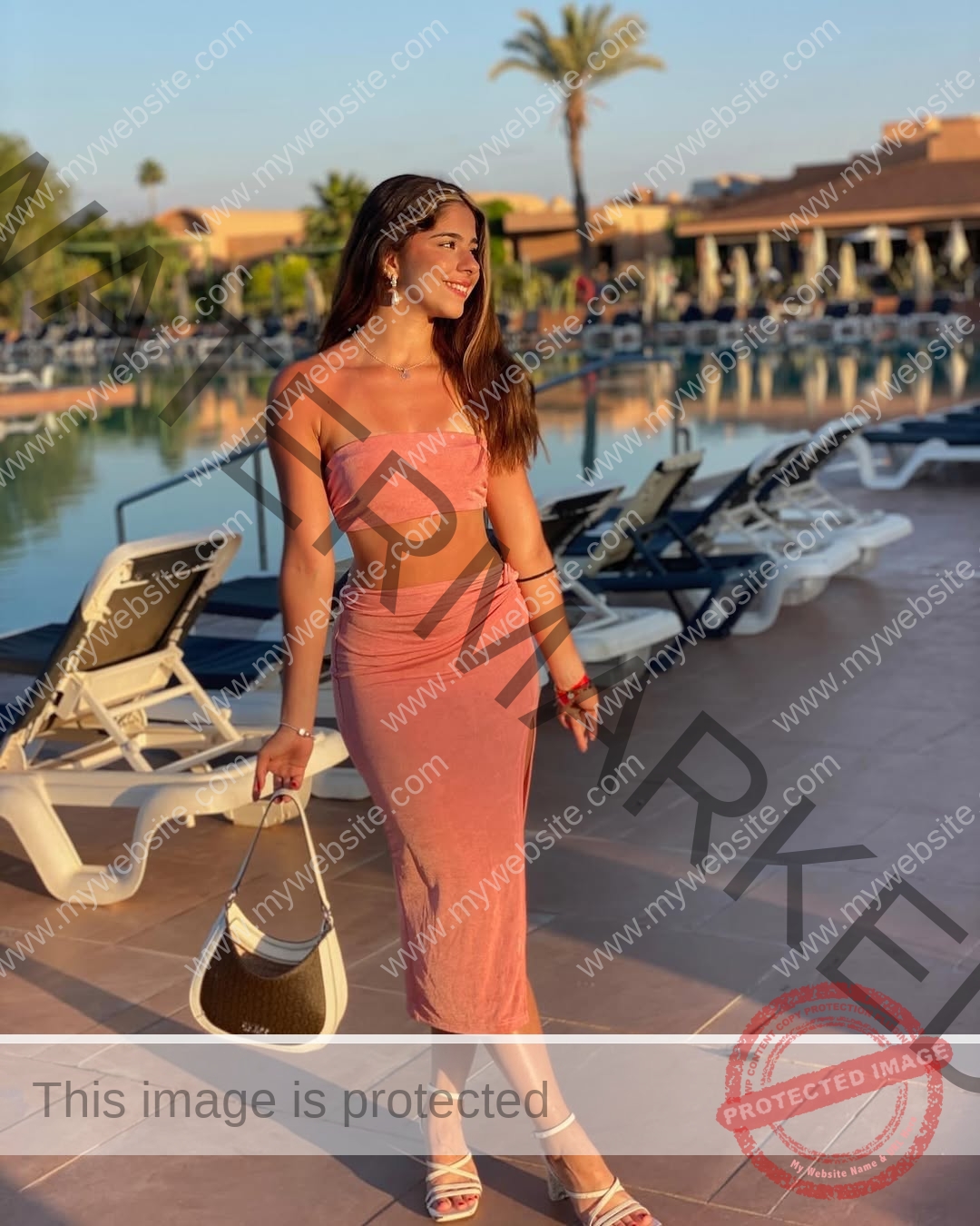 emma-deana-spain-emmadeana-15430 Emma Deana, track and field star from Spain, stands smiling by a pool in a pink outfit at sunset, palm trees and resort behind.