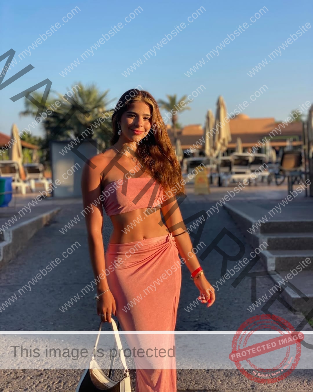 emma-deana-spain-emmadeana-15431 Emma Deana, track and field star from Spain, stands smiling outdoors in a pink strapless top and skirt, holding a black and white bag.