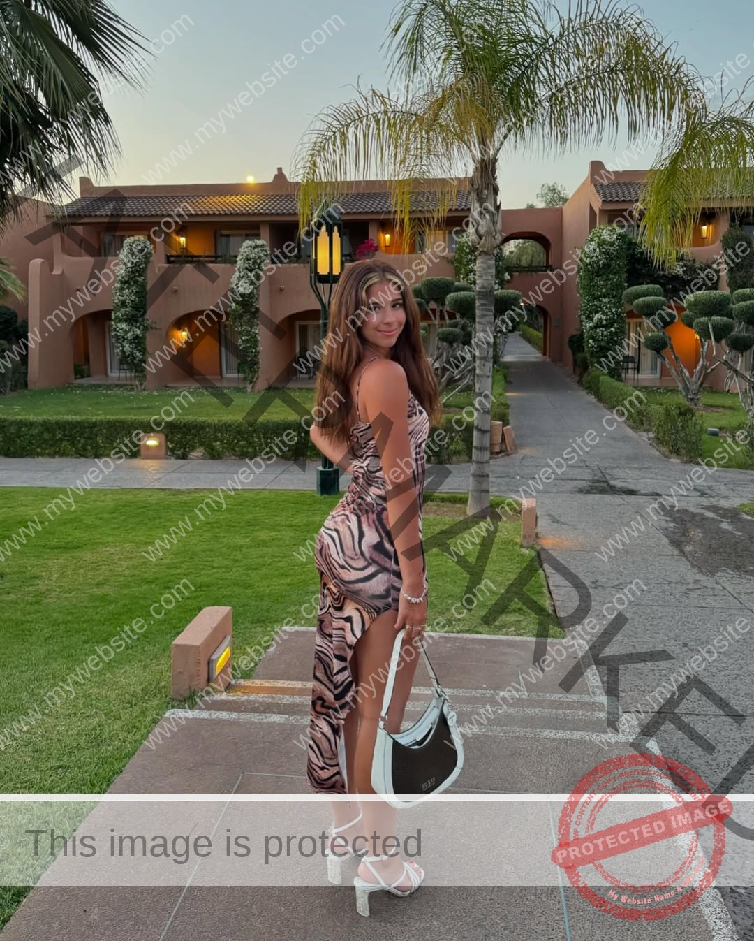 emma-deana-spain-emmadeana-15436 Emma Deana, track and field star from Spain, stands on a walkway in a patterned dress and white heels, holding a white handbag.