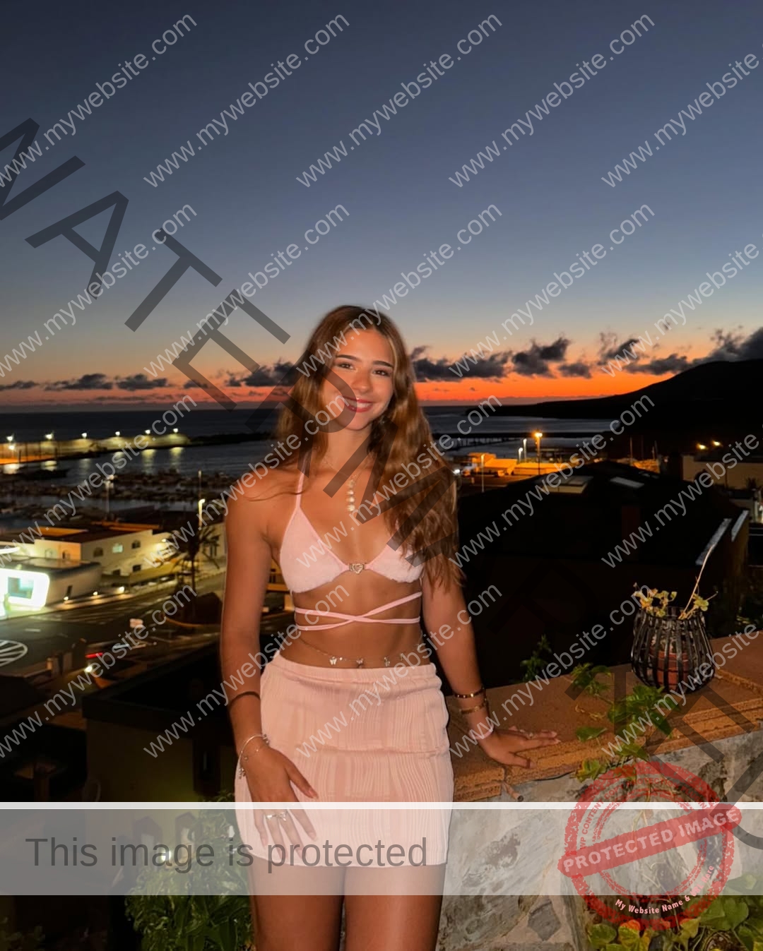emma-deana-spain-emmadeana-15439 Emma Deana, track and field star from Spain, with long brown hair in a pink crop top, smiles on a terrace at sunset by the ocean.