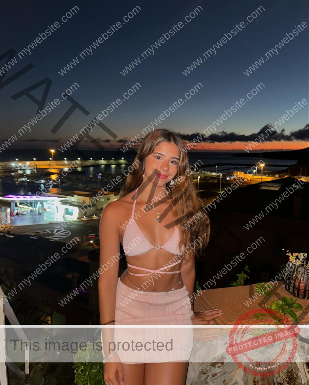 emma-deana-spain-emmadeana-15440 Emma Deana, track and field star from Spain, poses at sunset in a pink crop top and skirt before city lights and a dramatic sky.