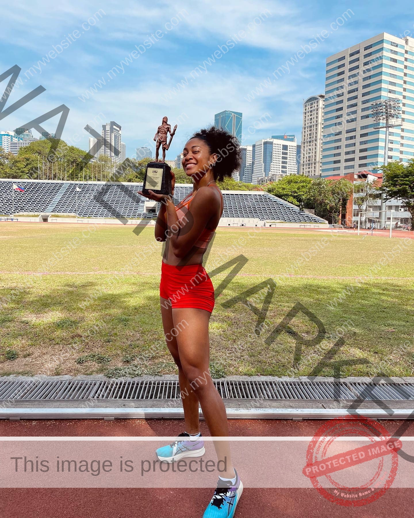 kristina-knott-philippines-knottyourcheese-x-18425 Kristina Knott, track and field star from Philippines, smiles in a red sports outfit holding a trophy on a track with city skyline.