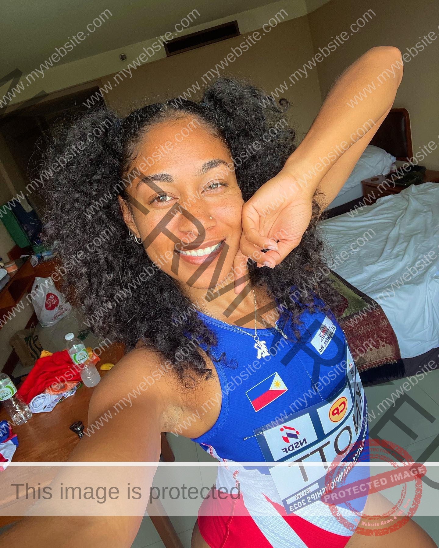 kristina-knott-philippines-knottyourcheese-x-18447 Kristina Knott, track and field star from Philippines, smiles for a selfie in a hotel room wearing blue athletic gear with flags.